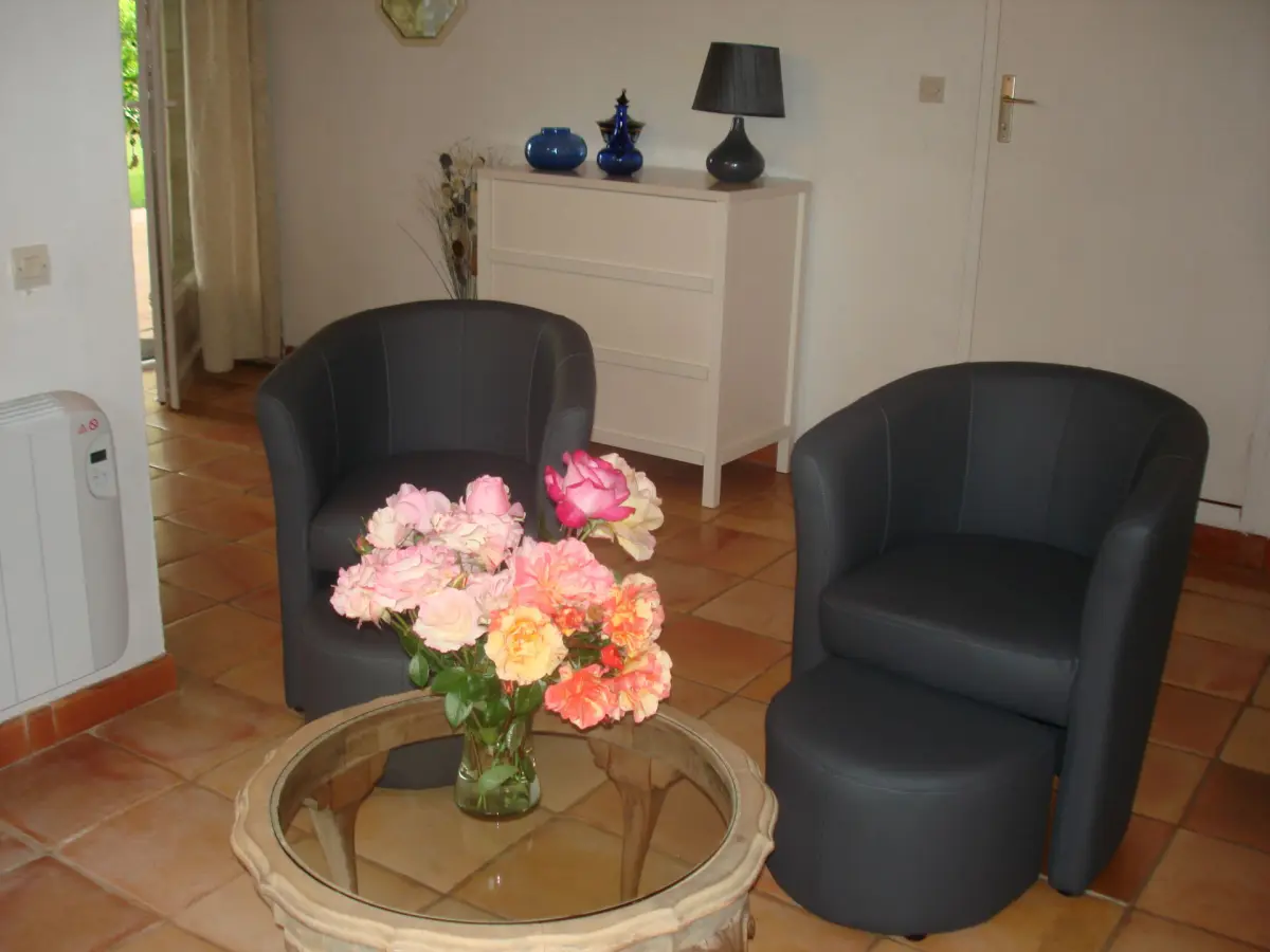 gite-coin-salon Vue du salon avec l'entrée du gîte, une commode, deux fauteuils et une table ronde avec un bouquet de fleurs du jardin