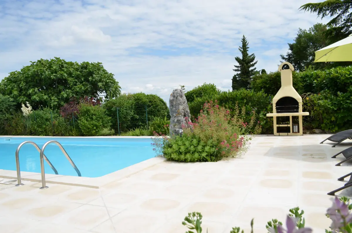 piscine-du-gite-cote-plage Vue partielle de la piscine depuis le portail d'entée avec l'échelle de bain, le barbecue, transats, parasol et la végétation tout autour